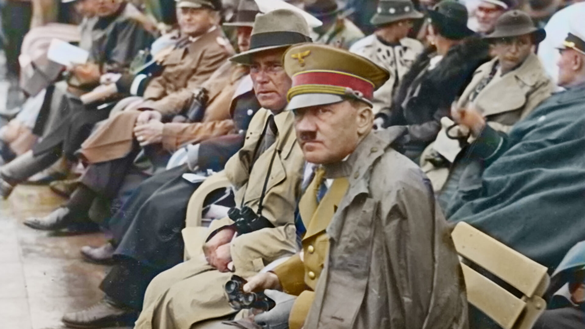 Group of men in military uniforms and civilians seated on folding chairs along a street during an outdoor event; a vintage color photo.