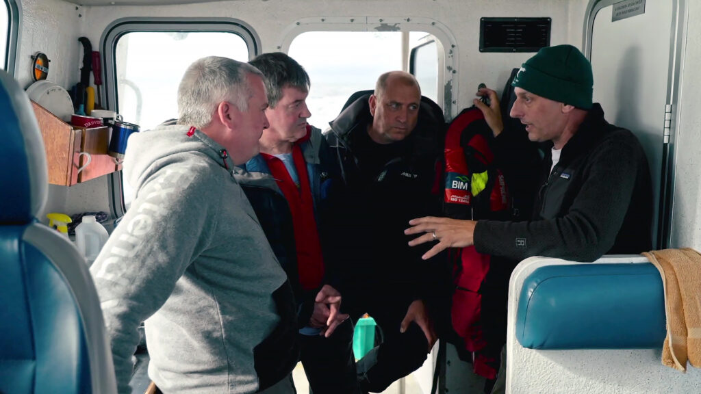 uboats_ep1_00135 - viasathistory.eu Four men in jackets inside a small boat cabin, looking and talking closely. Outside windows show a sea scene.